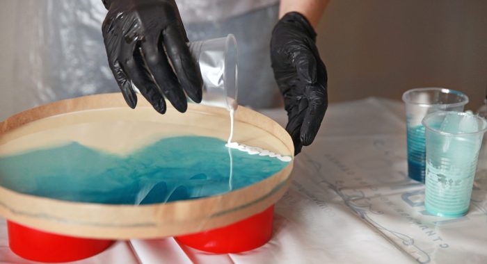 A girl artist in black gloves pours epoxy resin on a round surface and makes an artistic picture of the sea . Master class, hobby.